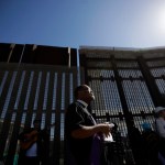 In this April 3, 2017 image, Reverend Guy A. Leemhuis, center, of the Holy Faith Episcopal Church of Los Angeles, leads a song in front of the border fence separating Tijuana, Mexico, from San Diego, in San Diego. With bids due Tuesday, April 4, 2017, on the first design contracts, companies are preparing for the worst if they get the potentially lucrative but controversial job. Four to 10 bidders are expected to be asked to build prototypes on a roughly quarter-mile (400-meter) strip of federally-owned land in San Diego, according to a U.S. official with knowledge of the plans who spoke on condition of anonymity because they haven't been made public. The land extends up to 120 feet (37 meters) from the border, raising the possibility of protests on both sides of the border.  (AP Photo/Gregory Bull)