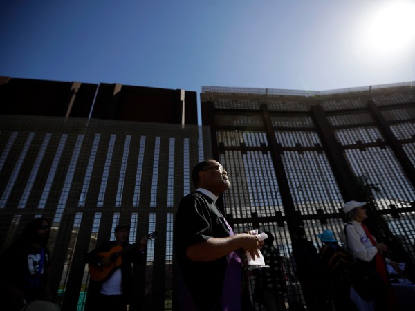 In this April 3, 2017 image, Reverend Guy A. Leemhuis, center, of the Holy Faith Episcopal Church of Los Angeles, leads a song in front of the border fence separating Tijuana, Mexico, from San Diego, in San Diego. With bids due Tuesday, April 4, 2017, on the first design contracts, companies are preparing for the worst if they get the potentially lucrative but controversial job. Four to 10 bidders are expected to be asked to build prototypes on a roughly quarter-mile (400-meter) strip of federally-owned land in San Diego, according to a U.S. official with knowledge of the plans who spoke on condition of anonymity because they haven't been made public. The land extends up to 120 feet (37 meters) from the border, raising the possibility of protests on both sides of the border.  (AP Photo/Gregory Bull)