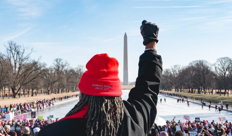 A woman stands on the steps of the Lincoln Memorial, facing the crowd and the Washington Monument, holding her fist up, and wearing a knitted cap with "Women's March Jan. 20, 2018" imprinted on it. People gathered around the reflecting pool for the "Women's March on Washington 2018", on Saturday, January 20, 2018. (Photo by Cheriss May) (Photo by Cheriss May/NurPhoto)