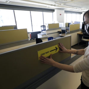 HOLD FOR STORY BY KELVIN CHAN — In this Thursday, May  7, 2020 photo, Interior Designer Stephanie Jones at the design firm Bergmeyer, applies a safe distancing reminder indicating one way foot traffic to a cubicle at the firms offices, in Boston. Out of concern for the coronavirus Bergmeyer is restructuring the way its workspace is used, including reinstalling dividers on 85 desks at its Boston office that had been removed over the years. (AP Photo/Steven Senne)