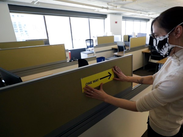 HOLD FOR STORY BY KELVIN CHAN — In this Thursday, May  7, 2020 photo, Interior Designer Stephanie Jones at the design firm Bergmeyer, applies a safe distancing reminder indicating one way foot traffic to a cubicle at the firms offices, in Boston. Out of concern for the coronavirus Bergmeyer is restructuring the way its workspace is used, including reinstalling dividers on 85 desks at its Boston office that had been removed over the years. (AP Photo/Steven Senne)