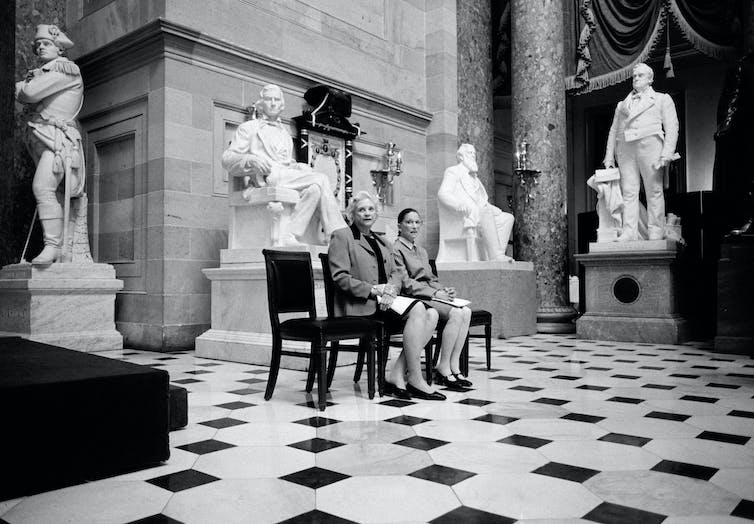 Two women seated in a large, marble-floored hall, surrounded by large statues of men.