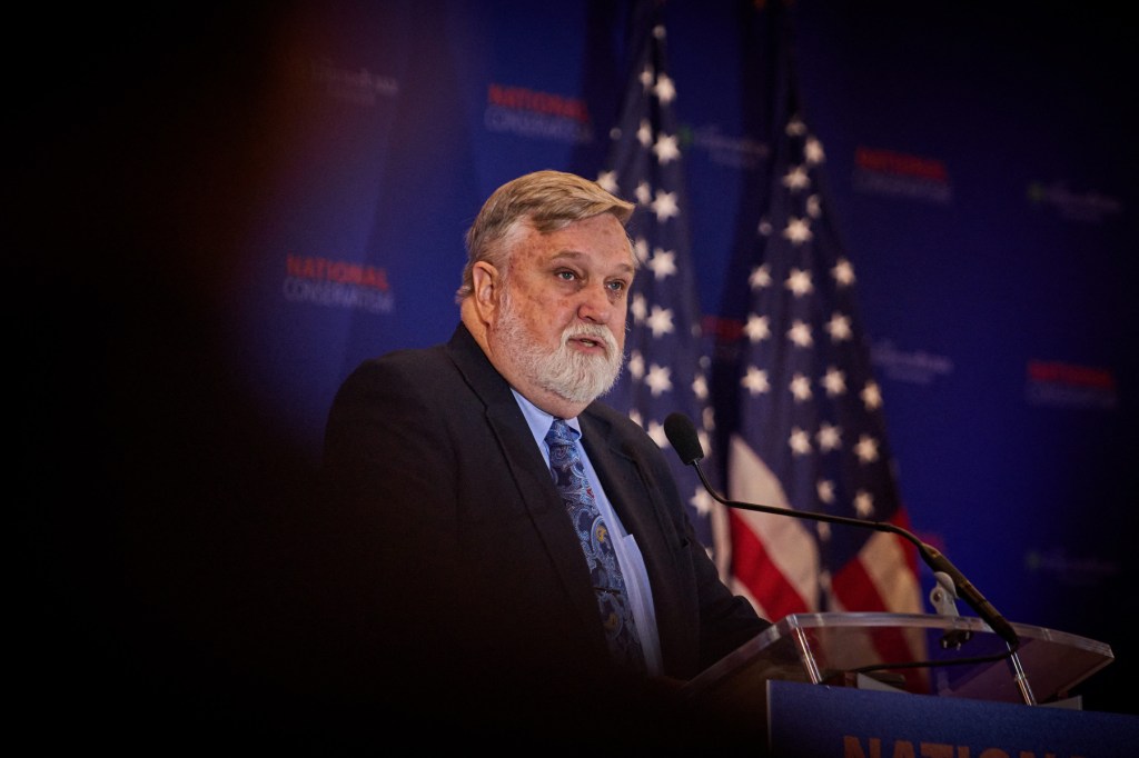 A gray-haired man speaks at a podium with an American flag behind him.