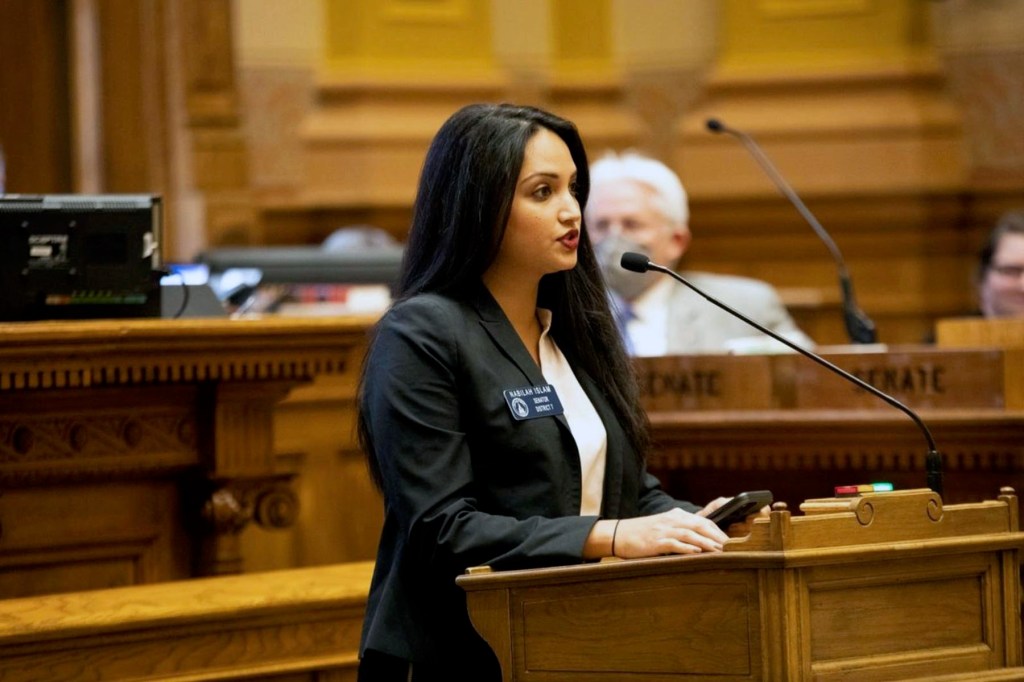 Nabilah Parkes stands at a desk on the Georgia Senate floor, speaking into a microphone with other lawmakers seated behind her.